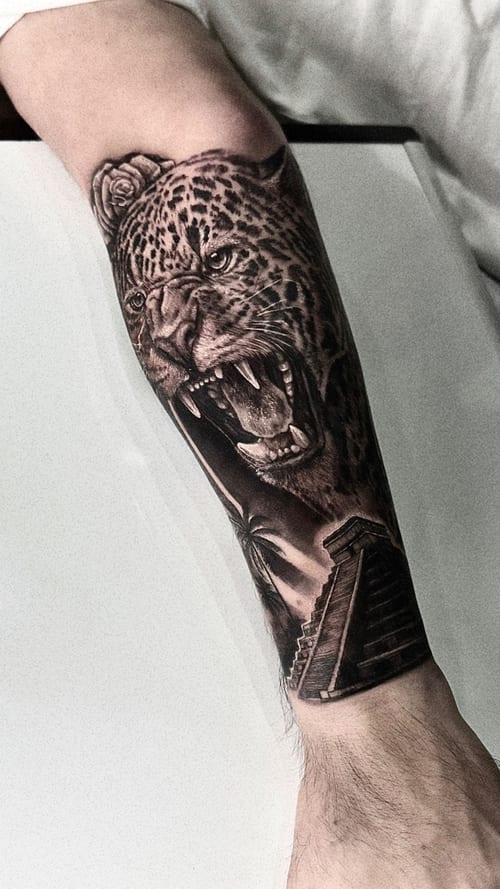 A detailed black and grey realism tattoo of a jaguar with an open mouth on the forearm, featuring a pyramid in the background — by INKEDBy_G, los angeles