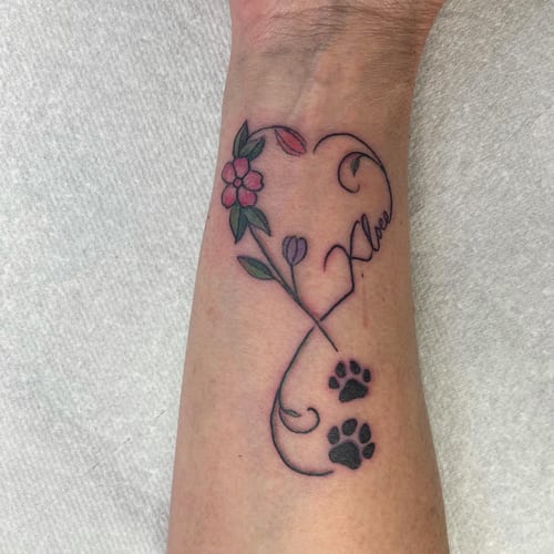 A delicate minimalist tattoo on the wrist featuring a flower and paw prints with fine line script — by Sandra Price, los angeles