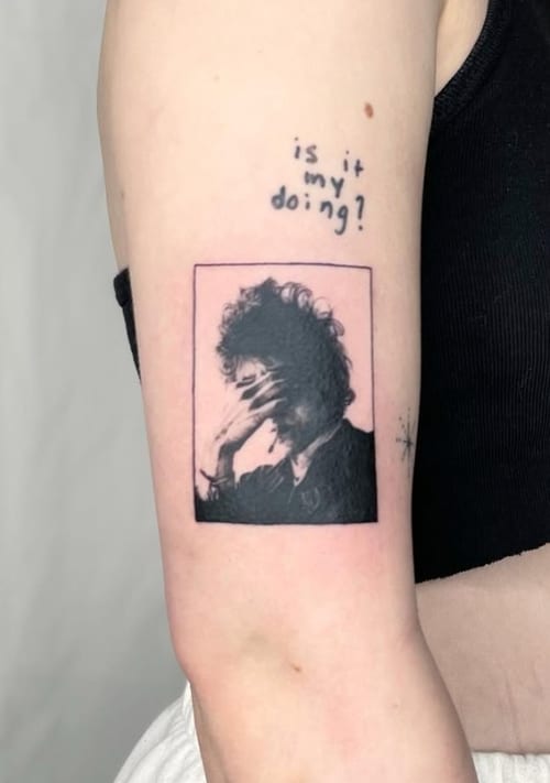 A black and grey portrait tattoo on the upper arm featuring a person with their hand covering their face, accompanied by the text 'is it my doing?' — by Level tattoo, los angeles