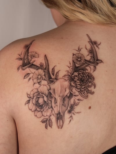 A black and grey tattoo of a deer skull surrounded by flowers on the back, featuring delicate and ornate illustrative details — by Melanie Benoit, los angeles