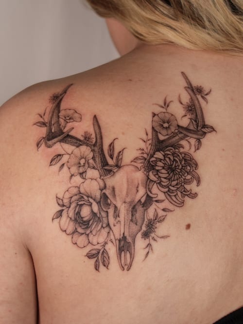 A black and grey tattoo of a deer skull surrounded by flowers on the back, featuring delicate and ornate illustrative details — by Melanie Benoit, los angeles