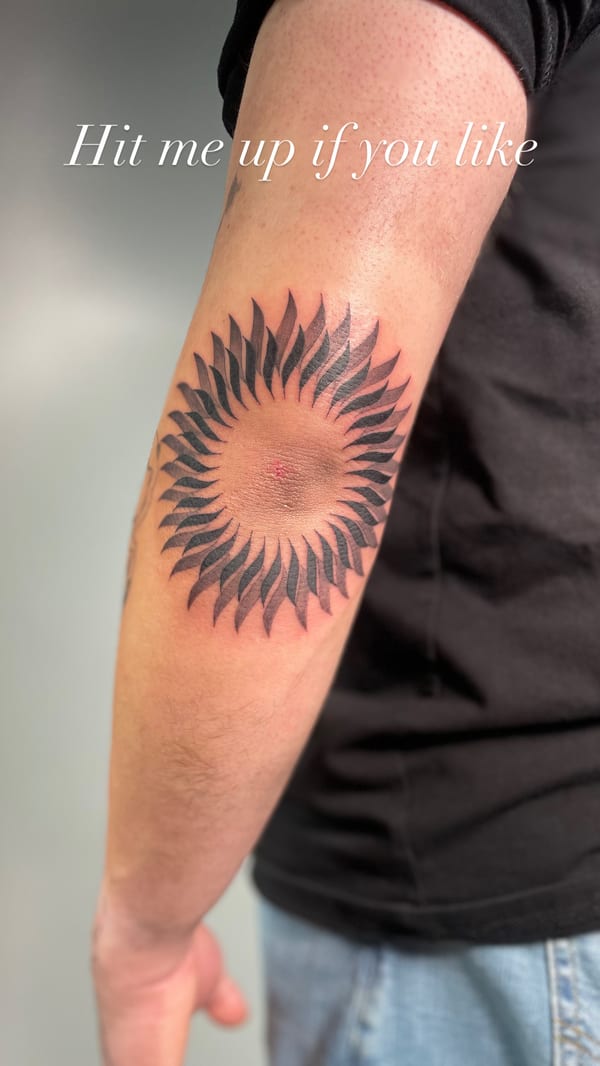 geometric blackwork sun tattoo with radiating blades wrapping around the elbow — by brian kies, los angeles