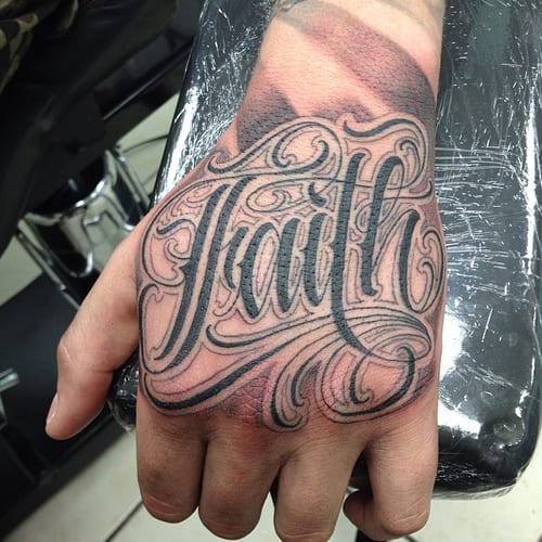 A bold black and grey script lettering tattoo of the word 'faith' on the hand — by Johnny Ayala, los angeles