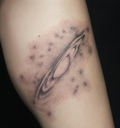 A black and grey abstract depiction of a galaxy on the thigh, featuring a central spiral with surrounding dots resembling stars — by Samantha Wilks, los angeles