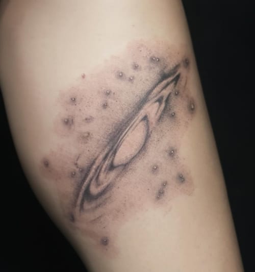 A black and grey abstract depiction of a galaxy on the thigh, featuring a central spiral with surrounding dots resembling stars — by Samantha Wilks, los angeles