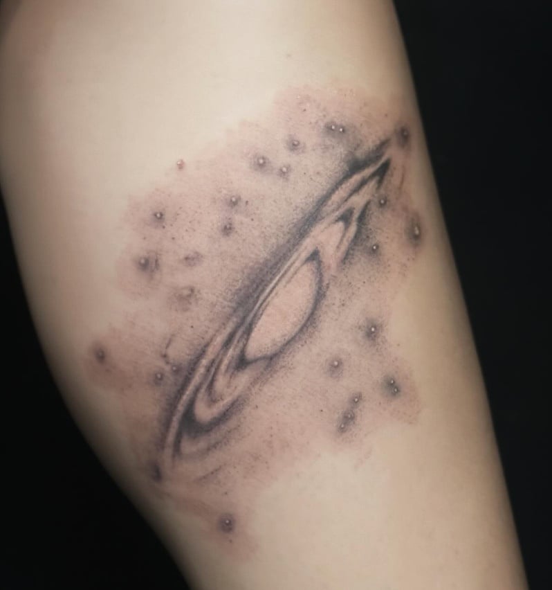 A black and grey abstract depiction of a galaxy on the thigh, featuring a central spiral with surrounding dots resembling stars — by Samantha Wilks, los angeles
