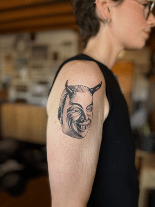 A black and grey portrait of a demonic face with horns on the upper arm, featuring bold lines and shading — by Dylan Aiello, los angeles