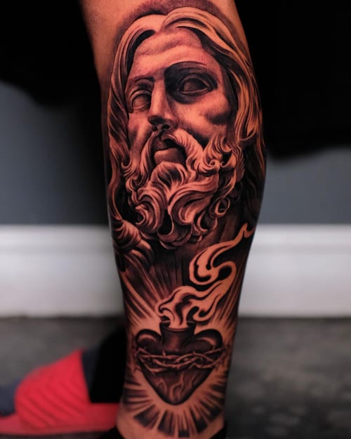 A detailed black and grey tattoo of Jesus Christ with a Sacred Heart on the calf, featuring realistic shading and spiritual symbolism — by Johnny Ayala, los angeles