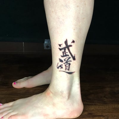 A bold black and grey kanji script tattoo on the ankle, featuring strong lettering in a blackwork style — by Sandra Price, los angeles