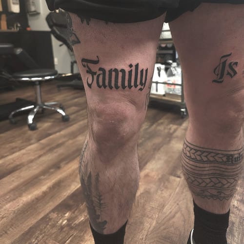 A bold black and grey script lettering tattoo on the thigh, featuring the word 'Family' in a classic font — by Miguel ANGELO, los angeles