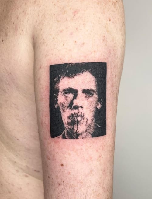 A black and grey portrait tattoo on the upper arm, featuring a stylized face with a square border and a distressed effect — by Level tattoo, los angeles