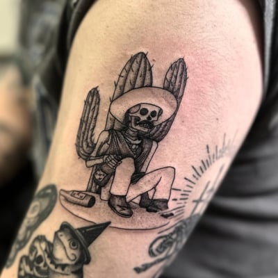 A black and grey tattoo of a skeleton wearing a sombrero, sitting with a cactus on the upper arm — by Maddie Ramirez, los angeles