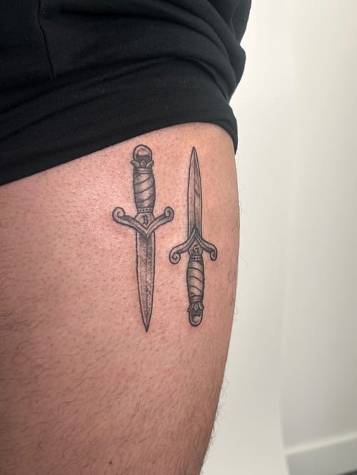 Two minimalist blackwork daggers on the thigh, featuring simple linework and shading — by Brian Kies, los angeles