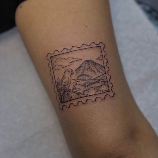 A minimalist black and grey tattoo of a parrot and mountain inside a stamp frame on the arm — by Maddie Ramirez, palm desert, ca