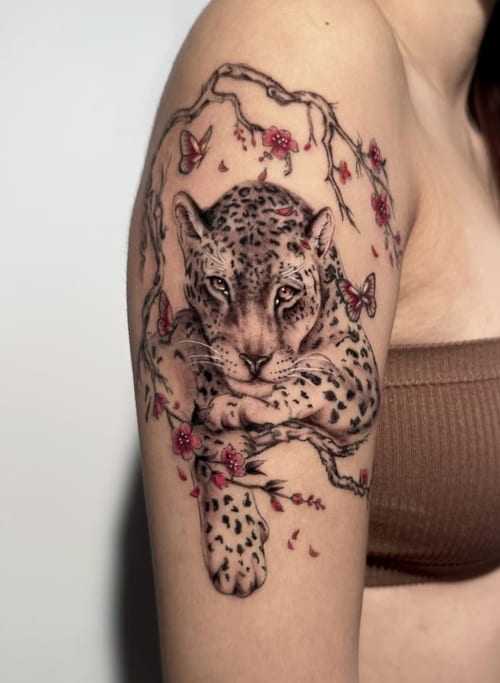 A realistic tattoo of a leopard resting on a branch with butterflies and flowers, featuring fine line work and a mix of color and black ink on the upper arm — by Shelby Bianco, los angeles
