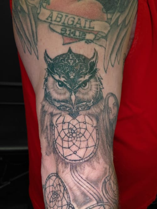 A black and grey tattoo of an owl with ornamental patterns and a dreamcatcher on the upper arm — by anoetic.designs.ultd, los angeles