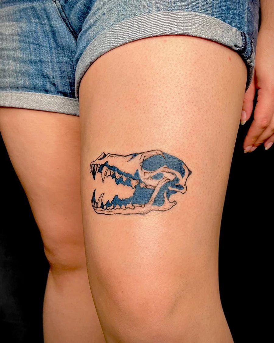 A graphic dinosaur skull tattoo on the thigh with black and blue ink, featuring bold outlines and illustrative style — by Dylan Aiello, los angeles