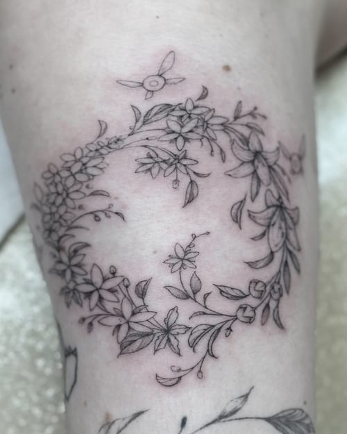 A delicate black and grey floral wreath tattoo on the thigh, featuring fine line botanical elements — by Melanie Benoit, los angeles