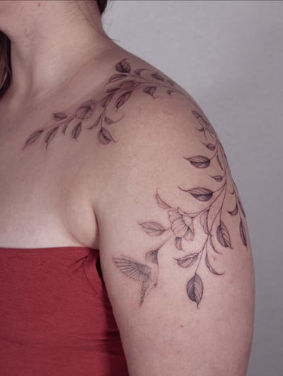 A delicate black and grey tattoo of a hummingbird with floral branches on the shoulder, featuring fine line work and botanical elements — by Melanie Benoit, los angeles