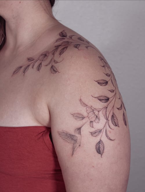A delicate black and grey tattoo of a hummingbird with floral branches on the shoulder, featuring fine line work and botanical elements — by Melanie Benoit, los angeles