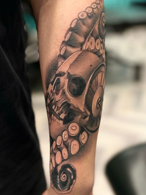 A black and grey tattoo on the forearm featuring a realistic skull entwined with octopus tentacles, showcasing detailed shading and texture — by Josue Bravo, los angeles
