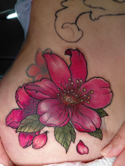 A vibrant neo-traditional floral tattoo on the hip, featuring large pink flowers with green leaves in a colorful style — by anoetic.designs.ultd, los angeles