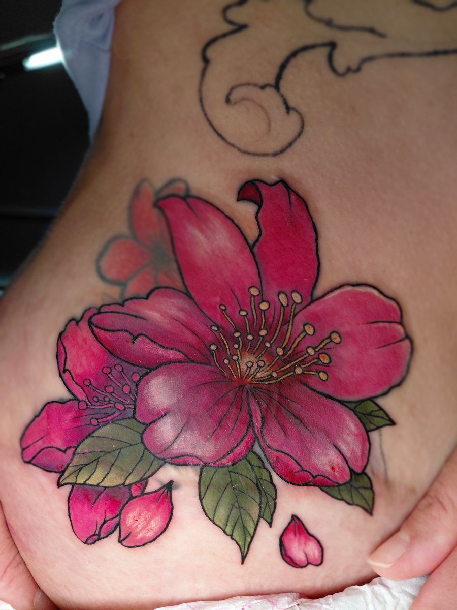A vibrant neo-traditional floral tattoo on the hip, featuring large pink flowers with green leaves in a colorful style — by anoetic.designs.ultd, los angeles
