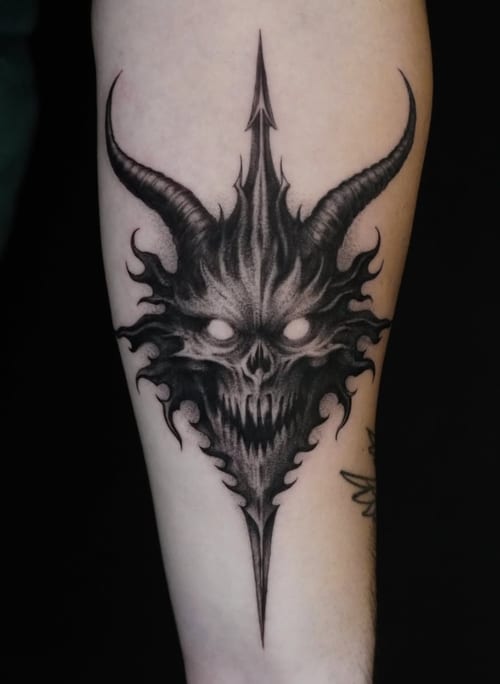 A black and grey tattoo of a demonic skull with horns on the forearm, featuring sharp lines and dark shading — by Jian Liang, los angeles