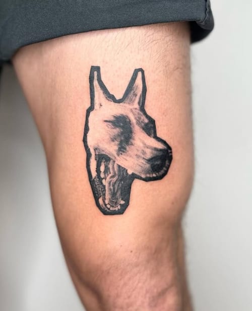 A black and grey surrealistic tattoo of a dog's head with a visible skeleton on the thigh, featuring bold outlines and shading — by Level tattoo, los angeles