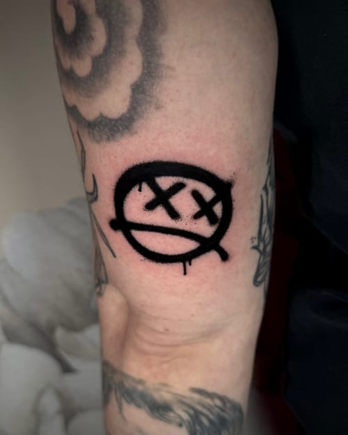A black graffiti-style face with crossed-out eyes on the upper arm, featuring bold lines and a simplistic design — by emily, los angeles