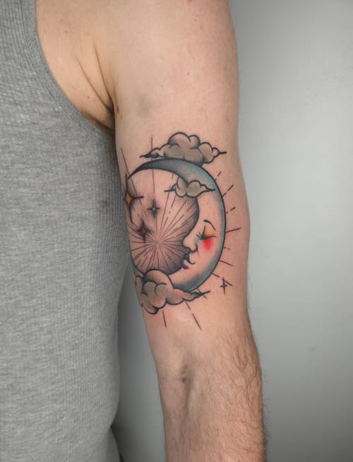 A colorful neo-traditional tattoo on the upper arm depicting a crescent moon with a face surrounded by clouds and stars, featuring bold outlines and vibrant shading — by ashynucklestattoo, los angeles