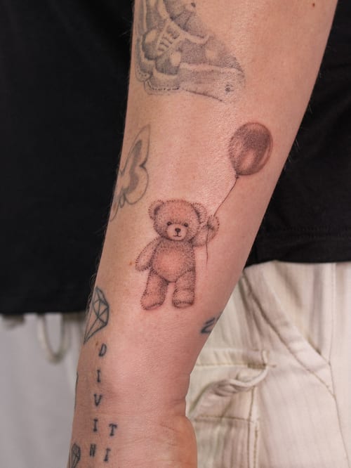 A delicate fine line tattoo of a teddy bear holding a balloon on the forearm, done in black and grey ink — by Melanie Benoit, los angeles