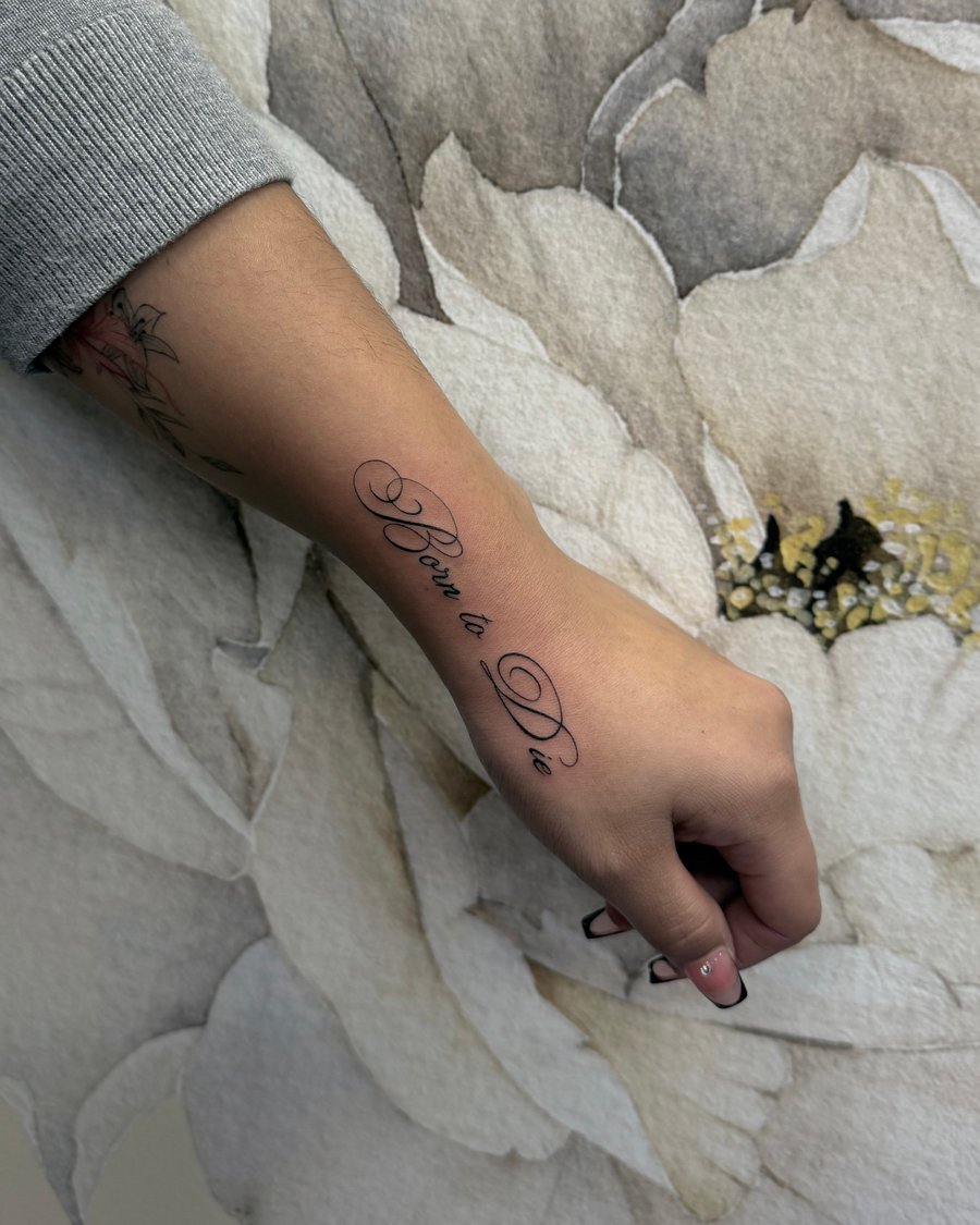 A black and grey script tattoo on the forearm with elegant lettering spelling 'Born to Die' — by emily, los angeles