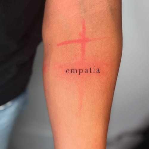 A minimalist tattoo of the word 'empatía' with a red cross on the forearm, combining lettering and simple line art — by zealous, los angeles