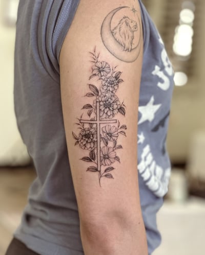 A delicate black and grey tattoo of a cross intertwined with flowers on the upper arm, done in a fine line style — by Melanie Benoit, los angeles
