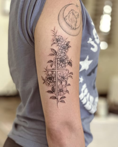 A delicate black and grey tattoo of a cross intertwined with flowers on the upper arm, done in a fine line style — by Melanie Benoit, los angeles