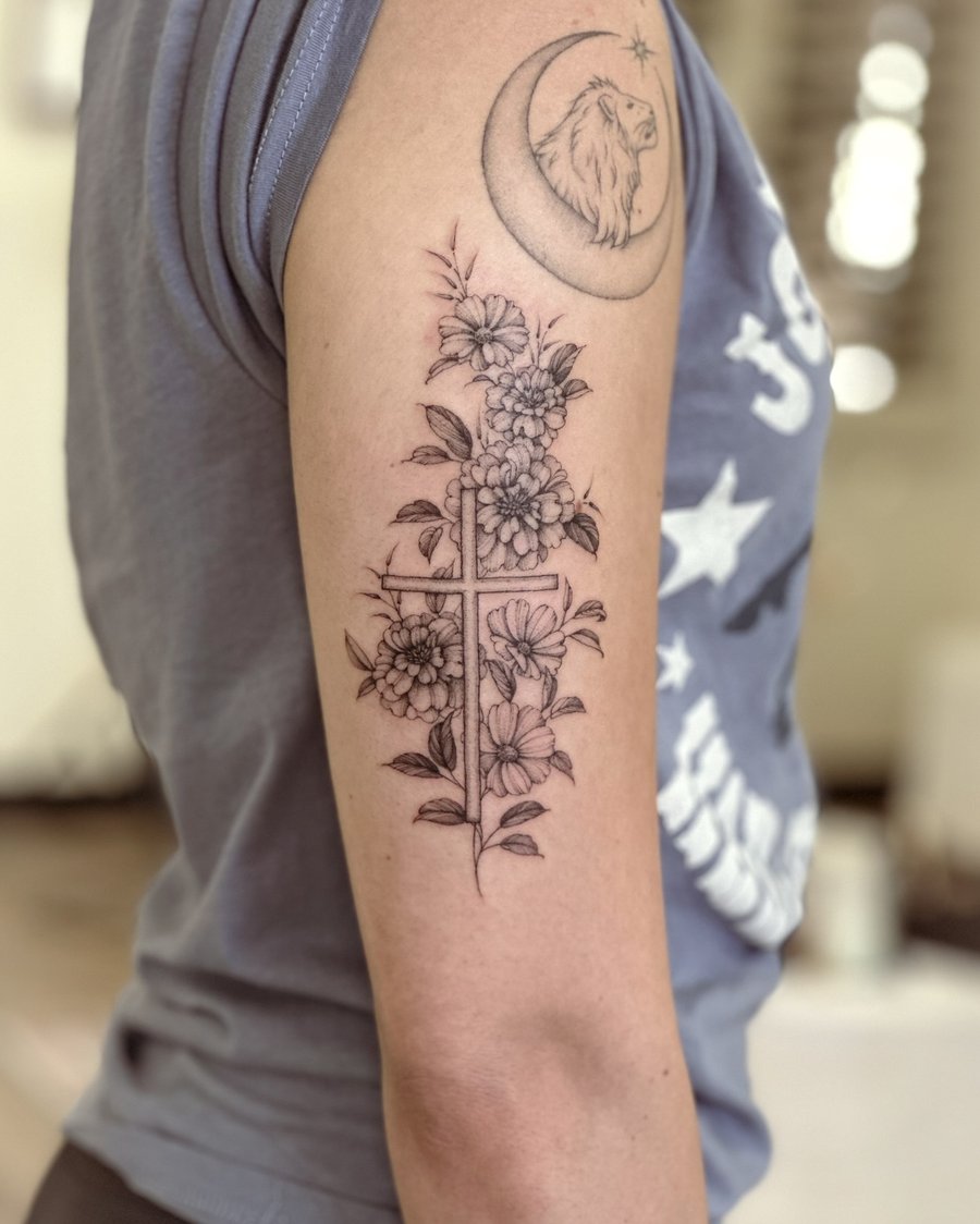 A delicate black and grey tattoo of a cross intertwined with flowers on the upper arm, done in a fine line style — by Melanie Benoit, los angeles