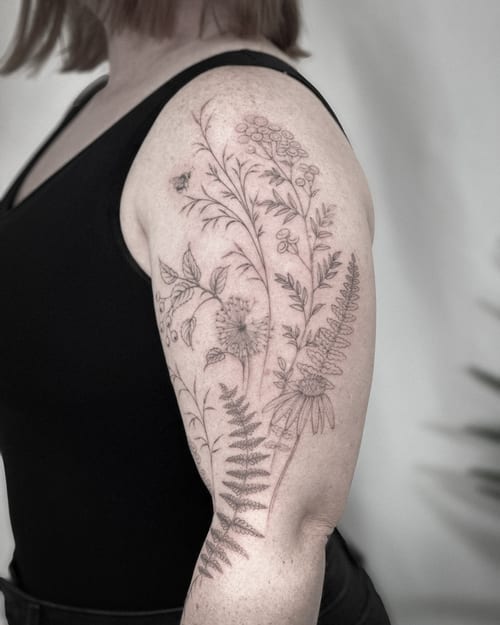 A delicate fine line botanical tattoo on the upper arm featuring various flowers and plants in black and grey — by Melanie Benoit, los angeles