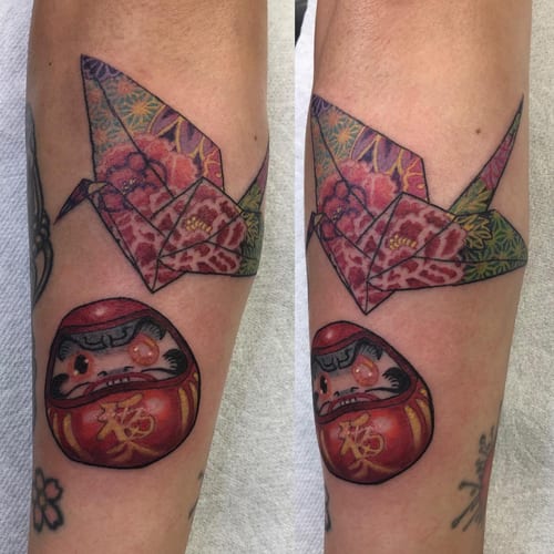 A colorful Japanese-style tattoo featuring an origami crane and a daruma doll on the forearm, showcasing intricate patterns and vibrant colors — by Sandra Price, los angeles