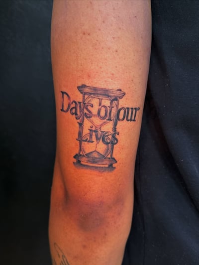 A black and grey hourglass with the text 'Days of our Lives' on the forearm, featuring bold script lettering — by Kiera Garvin, los angeles
