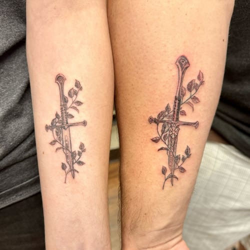 A delicate black and grey tattoo of a sword entwined with vines on the forearm, done in a fine line style — by Sandra Price, los angeles