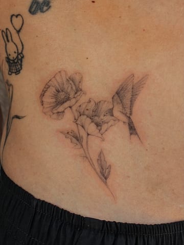 A delicate black and grey fine line tattoo of a hummingbird and flowers on the rib — by Melanie Benoit, los angeles