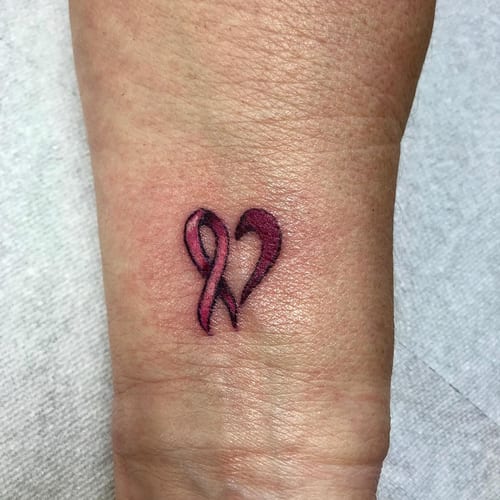 A small heart-shaped pink ribbon tattoo on the wrist, done in a minimalist and fine line style — by Sandra Price, los angeles