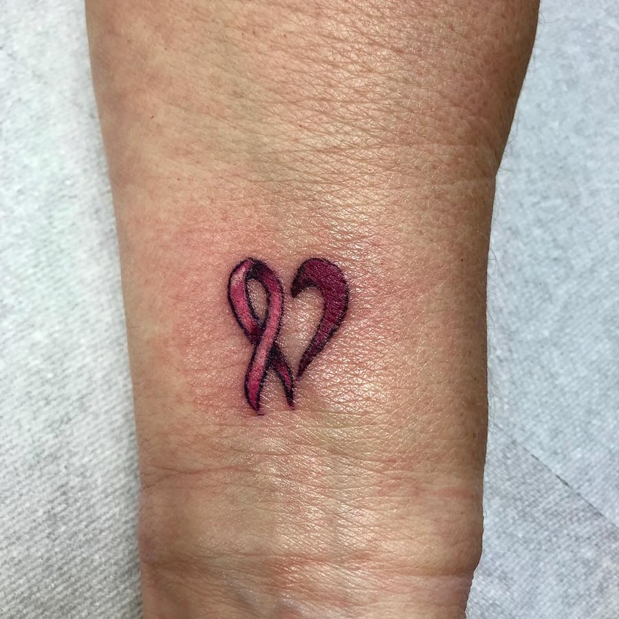 A small heart-shaped pink ribbon tattoo on the wrist, done in a minimalist and fine line style — by Sandra Price, los angeles
