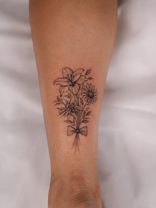 A delicate black and grey fine line flower bouquet tattoo on the forearm, featuring detailed floral elements tied with a bow — by Melanie Benoit, los angeles