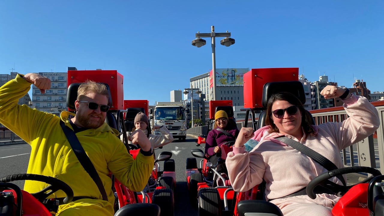 Adam and Lindsay smiling and waving while driving go karts through Tokyo