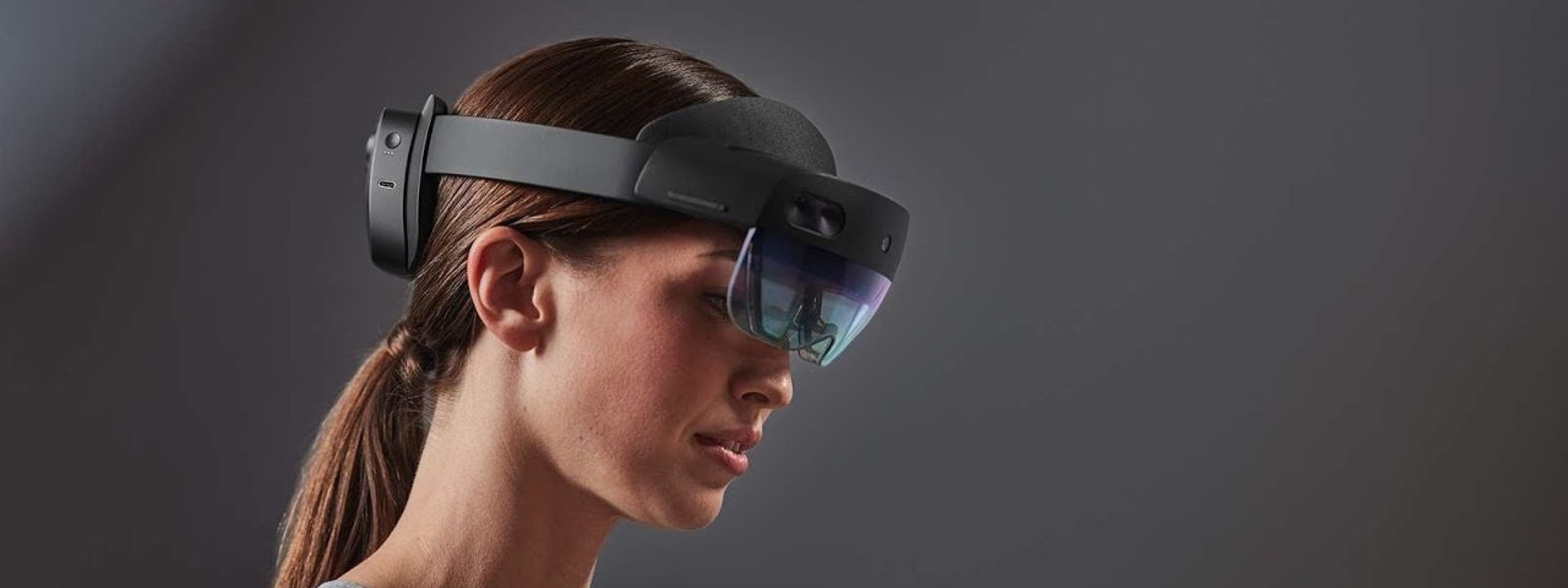 A woman with brown hair wearing a hololens on a dark grey background