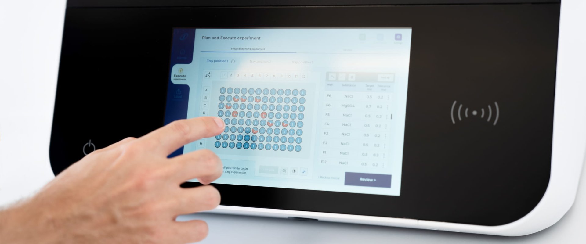 A hand pressing a blue Chemspeed touchscreen sitting on a black panel containing a UI showing a grid of dots some of which are blue and some are red