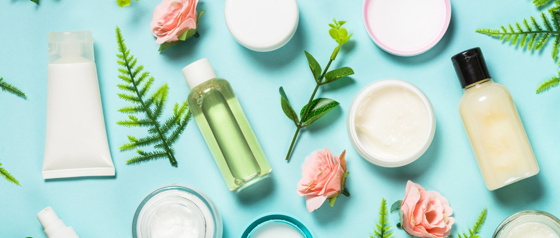 White creams and small clear plastic bottles containing clear and creamy liquid amongst pink rose heads and snippets of ferns on a teal background