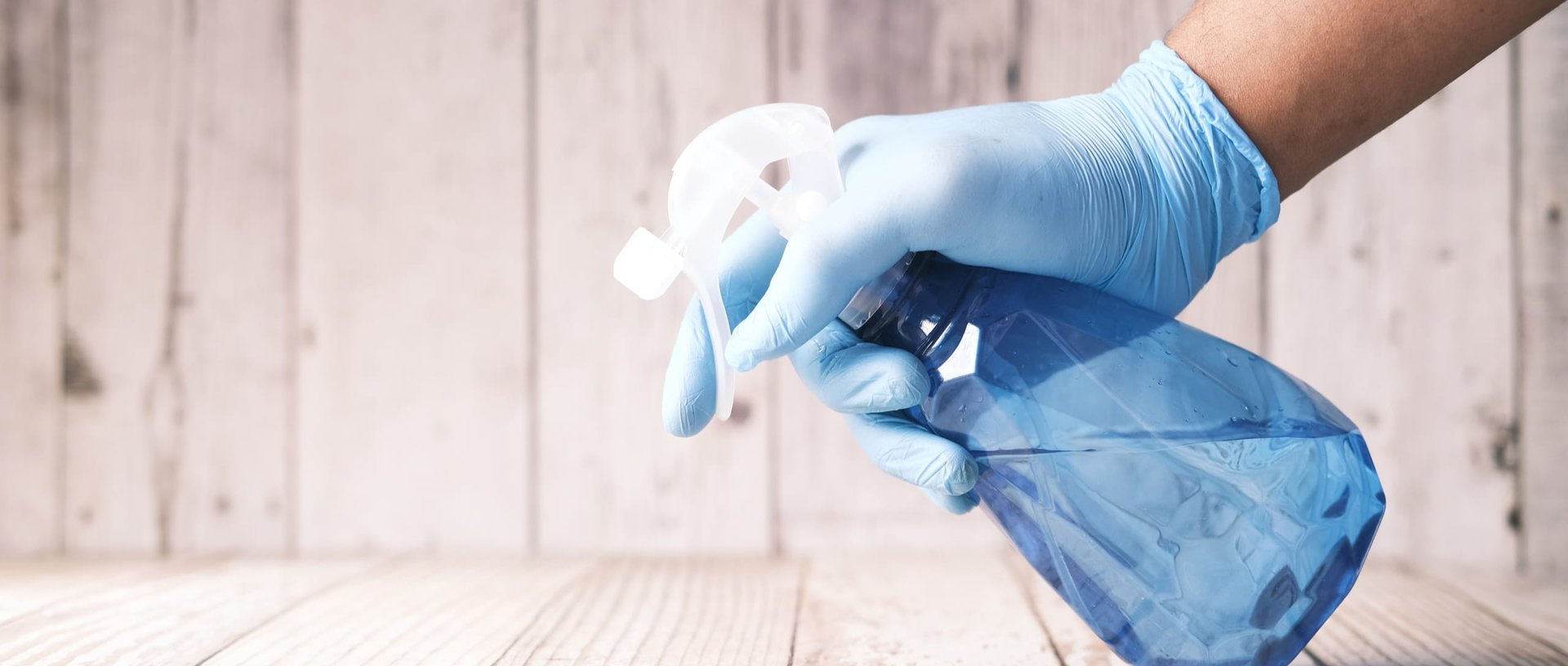 Hand in blue rubber gloves holding spray bottle cleaning table on wooden background